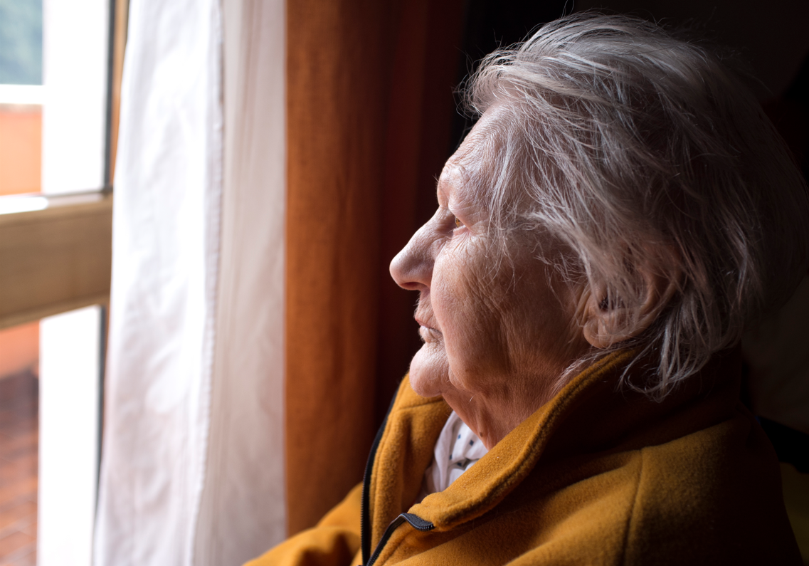 Profile shot of an elderly woman with short gray hair wearing a yellow fleece jacket, gazing thoughtfully out a bright window.